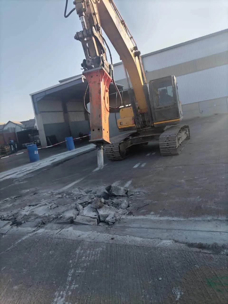 A tracked excavator fitted with a hydraulic breaker attachment, breaking concrete.
